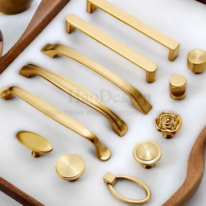 Può includere: Una collezione di ferramenta dorata per mobili, tra cui maniglie, pomelli e un anello. L'hardware presenta vari design, tra cui maniglie a barra, maniglie curve e un pomello a forma di rosa. Gli articoli sono disposti su una superficie bianca all'interno di un vassoio di legno.