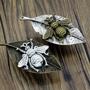 May include: Two silver and bronze bee charms on leaf-shaped pendants. The bees have detailed wings and bodies. The leaves have a textured surface.