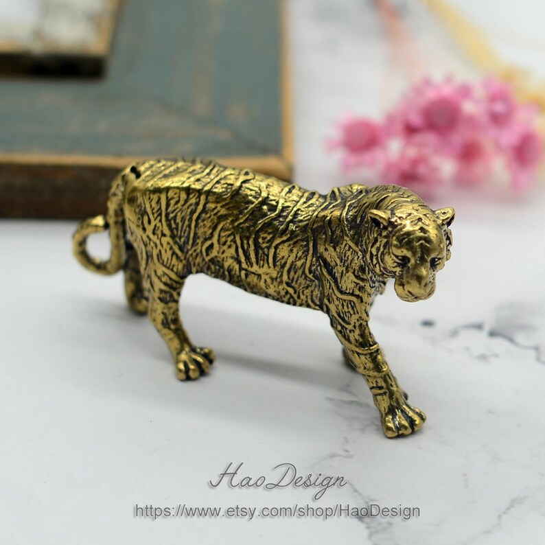 Pode incluir: Uma pequena figura de metal dourada de um tigre. O tigre est&aacute; em p&eacute; sobre suas quatro patas e tem uma superf&iacute;cie detalhada e texturizada.