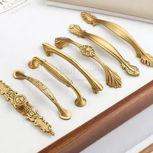 K&ouml;nnte beinhalten: Eine Sammlung verzierter, goldfarbener Schrankgriffe und -kn&ouml;pfe. Die Hardware weist verschiedene Designs auf, darunter Blumen- und Scrollmuster. Die Griffe sind auf einer wei&szlig;en Oberfl&auml;che mit einem Holzrahmen angeordnet und zeigen ihre dekorativen Details.