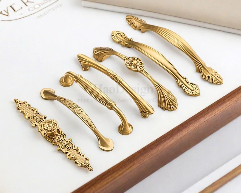 K&ouml;nnte beinhalten: Eine Sammlung verzierter, goldfarbener Schrankgriffe und -kn&ouml;pfe. Die Hardware weist verschiedene Designs auf, darunter Blumen- und Scrollmuster. Die Griffe sind auf einer wei&szlig;en Oberfl&auml;che mit einem Holzrahmen angeordnet und zeigen ihre dekorativen Details.