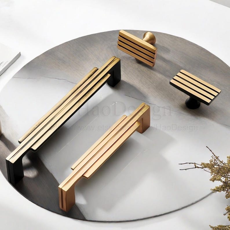 Op de afbeelding: Een verzameling gouden en zwarte kastgrepen en knoppen. De handgrepen hebben een lineair ontwerp met drie parallelle staven. De knoppen hebben een rechthoekige vorm en een ronde basis. De hardware wordt gepresenteerd op een marmeren oppervlak.