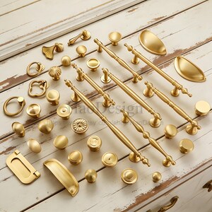 May include: A collection of gold-colored cabinet hardware, including knobs, pulls, and handles, arranged on a white wooden surface. The hardware features various designs, from simple round knobs to ornate pulls with decorative detailing. The image showcases a variety of styles.