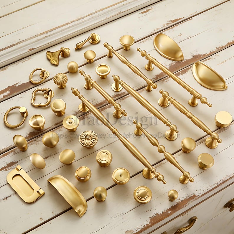 May include: A collection of gold-colored cabinet hardware, including knobs, pulls, and handles, arranged on a white wooden surface. The hardware features various designs, from simple round knobs to ornate pulls with decorative detailing. The image showcases a variety of styles.