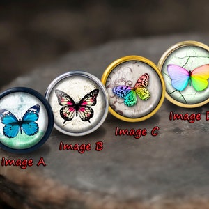 以下が含まれることがあります： 異なる蝶のデザインが施された丸いガラス製のカボションが4つあります。蝶は青、ピンク、虹色、そして多色です。カボションは、金、銀、黒の仕上げの金属製のセッティングにセットされています。