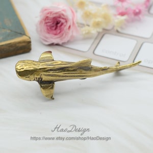 May include: A brass shark figurine with a detailed, textured surface. The figurine is positioned on a white surface.