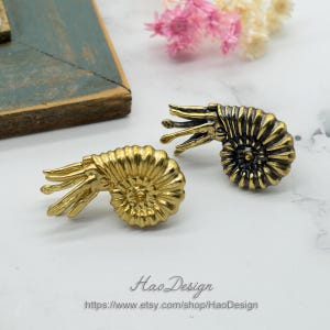May include: Two gold-toned ammonite shell earrings with intricate details. The shell on the left is solid gold, while the shell on the right has a black and gold finish.