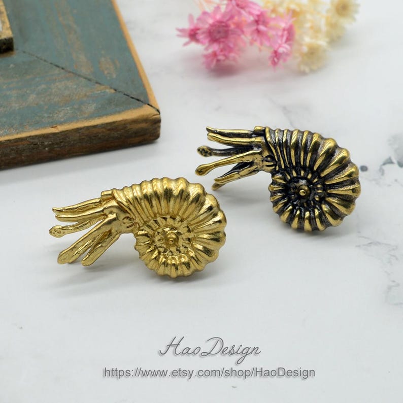 May include: Two gold-toned ammonite shell earrings with intricate details. The shell on the left is solid gold, while the shell on the right has a black and gold finish.