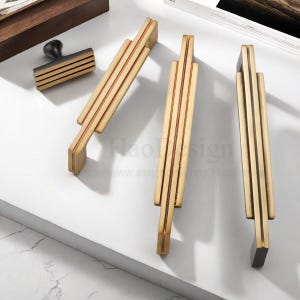 Op de afbeelding: Een verzameling goudkleurige kastbeslagen, waaronder drie lange handgrepen en een kleine knop. De handgrepen hebben een gelaagd ontwerp met verticale groeven, terwijl de knop rechthoekig is met horizontale groeven. De hardware wordt getoond op een wit oppervlak.