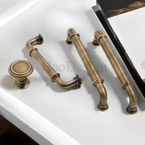 May include: An assortment of antique brass cabinet hardware, including two pull handles, a curved handle, and a round knob. The hardware has a vintage aesthetic with a warm, aged finish, perfect for kitchen or bathroom renovations.