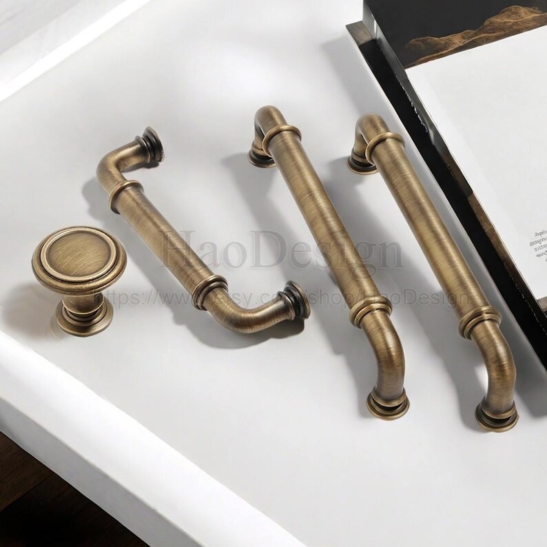 May include: An assortment of antique brass cabinet hardware, including two pull handles, a curved handle, and a round knob. The hardware has a vintage aesthetic with a warm, aged finish, perfect for kitchen or bathroom renovations.