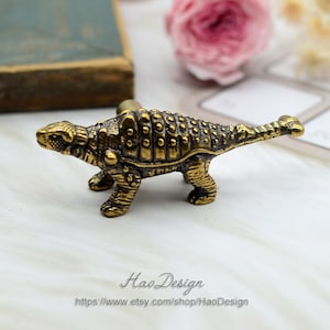 May include: A gold-toned Ankylosaurus dinosaur figurine with a textured back and a long tail. The figurine is a cabinet knob or drawer pull.