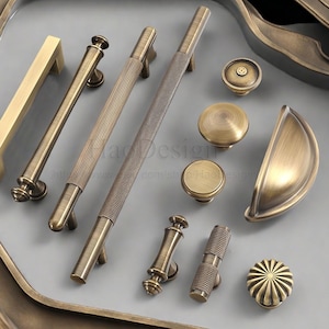 Könnte beinhalten: Eine Sammlung antiker Messing-Schrankbeschläge, darunter verschiedene Griffe, Knöpfe und ein Griff. Die Griffe weisen unterschiedliche Designs auf, wie gerippte und strukturierte Oberflächen. Die Beschläge sind auf einer hellgrauen Oberfläche angeordnet.