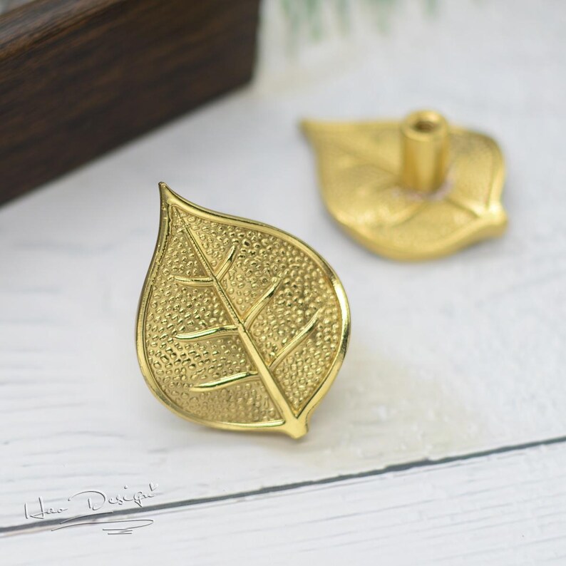 K&ouml;nnte beinhalten: Zwei goldene, blattf&ouml;rmige Schrankkn&ouml;pfe mit strukturierter Oberfl&auml;che. Die Kn&ouml;pfe haben ein kleines Loch in der Mitte zum Befestigen.