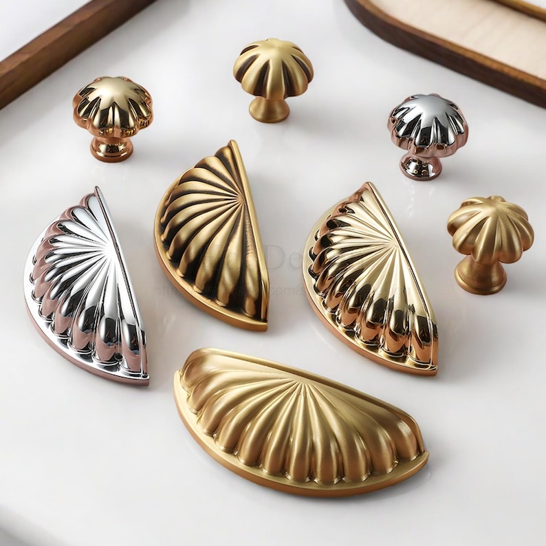 Może przedstawiać: Kolekcja dekoracyjnych okuć do szafek, w tym złote, srebrne i r&oacute;żowo-złote gałki i uchwyty. Gałki mają kształt grzyb&oacute;w, a uchwyty są p&oacute;łokrągłe ze wzorem wachlarza.