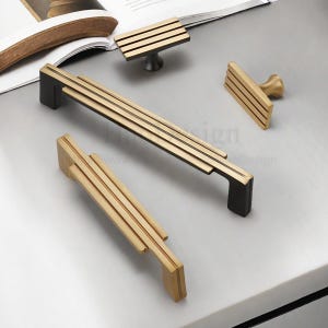 Op de afbeelding: Een verzameling gouden en zwarte kastbeslag, waaronder handgrepen en knoppen. De handgrepen hebben een lineair ontwerp met meerdere parallelle staven. De knoppen hebben een vergelijkbaar ontwerp. Het beslag wordt gepresenteerd op een lichtgrijze ondergrond.