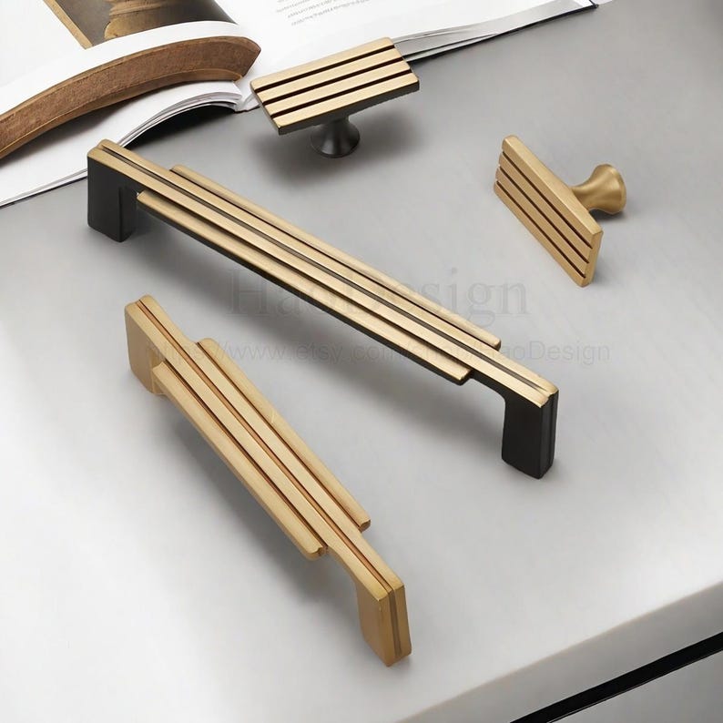 Op de afbeelding: Een verzameling gouden en zwarte kastbeslag, waaronder handgrepen en knoppen. De handgrepen hebben een lineair ontwerp met meerdere parallelle staven. De knoppen hebben een vergelijkbaar ontwerp. Het beslag wordt gepresenteerd op een lichtgrijze ondergrond.