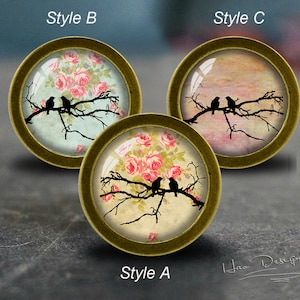 Op de afbeelding: Drie ronde, decoratieve, messingkleurige, glasachtige ornamenten met verschillende ontwerpen. De eerste ornament heeft een bloemenpatroon met roze rozen en een paar vogels op een tak. De tweede ornament heeft een bloemenpatroon met roze rozen en een paar vogels op een tak. De derde ornament heeft een aquarelachtige achtergrond met een paar vogels op een tak.