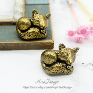 Può includere: Due statuette di volpe color bronzo in posizione rannicchiata mentre dormono. Le volpi hanno una pelliccia strutturata e piccole orecchie. Le statuette sono esposte su una superficie chiara. Il testo "Hao Design" e un indirizzo web sono visibili.