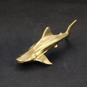 May include: A gold-colored metal shark figurine. The shark is depicted in a swimming pose with its fins spread out.