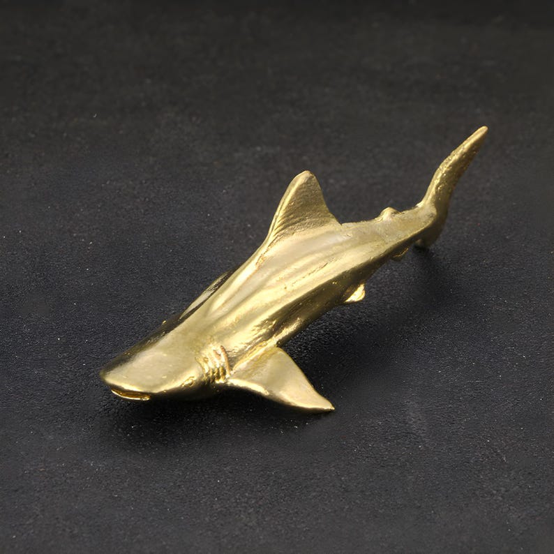 May include: A gold-colored metal shark figurine. The shark is depicted in a swimming pose with its fins spread out.