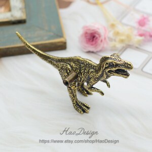 Pode incluir: Uma ma&ccedil;aneta de arm&aacute;rio ou puxador de gaveta em forma de dinossauro de cor dourada. O dinossauro est&aacute; em p&eacute; sobre suas quatro patas com a cabe&ccedil;a levantada e a boca ligeiramente aberta. O dinossauro tem uma superf&iacute;cie detalhada e texturizada.