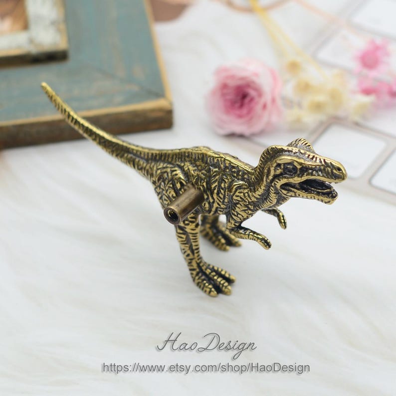 Pode incluir: Uma ma&ccedil;aneta de arm&aacute;rio ou puxador de gaveta em forma de dinossauro de cor dourada. O dinossauro est&aacute; em p&eacute; sobre suas quatro patas com a cabe&ccedil;a levantada e a boca ligeiramente aberta. O dinossauro tem uma superf&iacute;cie detalhada e texturizada.