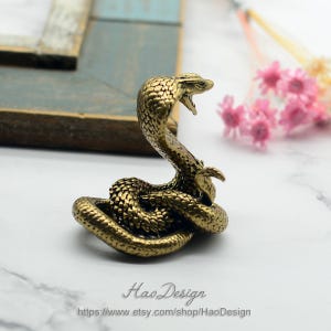 May include: A small, gold-coloured brass sculpture of a cobra with its hood raised, in a coiled position. The sculpture is detailed with scales and a textured surface.