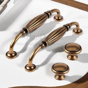 May include: Two antique bronze cabinet pulls and two matching knobs. The pulls have a curved handle and a ribbed center section. The knobs are round with a raised center. The hardware is displayed on a white surface.