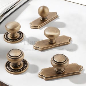 May include: A selection of antique bronze cabinet knobs and pulls. The knobs are spherical, and some feature decorative rope detailing. The pulls have a rectangular backplate with a rounded top and bottom. The hardware is displayed on a white surface.