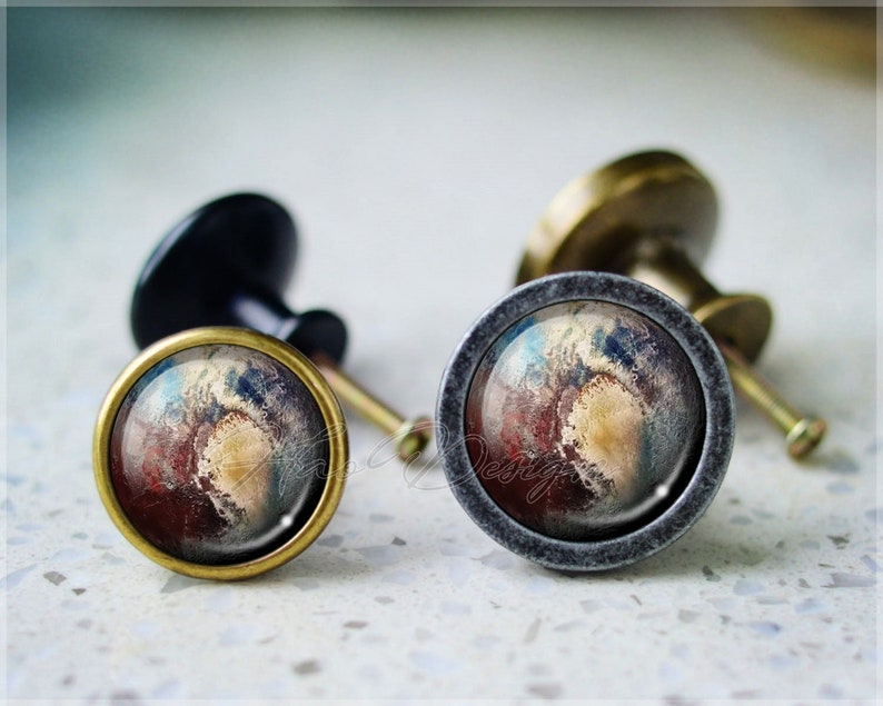 K&ouml;nnte beinhalten: Zwei dekorative Schrankkn&ouml;pfe mit einer runden Messingbasis und einem grauen Metallring. Die Kn&ouml;pfe zeigen ein farbenfrohes, abstraktes Design in Rot-, Blau- und Gelbt&ouml;nen.