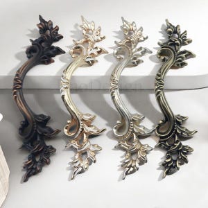 Pode incluir: Quatro puxadores de armário ornamentados em tons de bronze, ouro e prata. Cada puxador apresenta um design curvo com detalhes florais e de folhas. Os puxadores estão dispostos sobre uma superfície branca, mostrando seus detalhes decorativos.