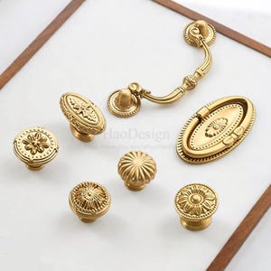 K&ouml;nnte beinhalten: Eine Sammlung antiker Messing-Schrankbeschl&auml;ge, darunter Kn&ouml;pfe und Griffe. Die Beschl&auml;ge weisen verzierte Blumen- und geometrische Designs auf. Die Teile sind auf einer wei&szlig;en Oberfl&auml;che mit einem Holzrahmen angeordnet.