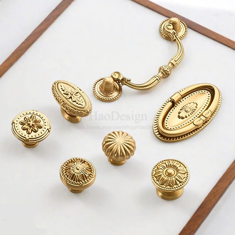 K&ouml;nnte beinhalten: Eine Sammlung antiker Messing-Schrankbeschl&auml;ge, darunter Kn&ouml;pfe und Griffe. Die Beschl&auml;ge weisen verzierte Blumen- und geometrische Designs auf. Die Teile sind auf einer wei&szlig;en Oberfl&auml;che mit einem Holzrahmen angeordnet.