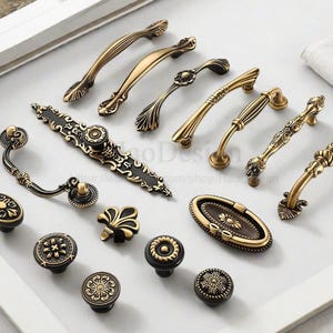 以下が含まれることがあります： 金と黒のアンティークスタイルのキャビネットハンドルとノブのコレクション。金具には、華やかな花柄とスクロールワークのデザインが施されています。商品は白い表面に配置されています。寸法はセンチメートルです。