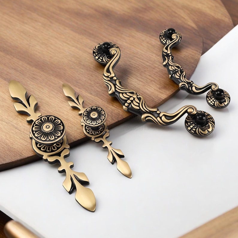 Op de afbeelding: Een assortiment antieke meubelbeslag. De handgrepen en knoppen hebben een goud- en zwarte afwerking, met sierlijke bloemen- en bladerenontwerpen. Het beslag wordt gepresenteerd op een houten oppervlak.