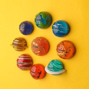 Könnte beinhalten: Ein Set aus neun bunten Magneten in Form von Planeten, darunter Erde, Neptun, Venus, Jupiter, Mars, Uranus, Saturn und die Sonne. Jeder Magnet hat eine Beschriftung mit dem Namen und ein einzigartiges Design.