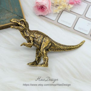 Pode incluir: Uma figurinha de dinossauro de metal dourado. O dinossauro est&aacute; em p&eacute; sobre uma superf&iacute;cie branca. O dinossauro est&aacute; olhando para o lado direito da imagem. O dinossauro tem uma cauda longa e uma cabe&ccedil;a pequena. O dinossauro &eacute; detalhado com escamas texturizadas.