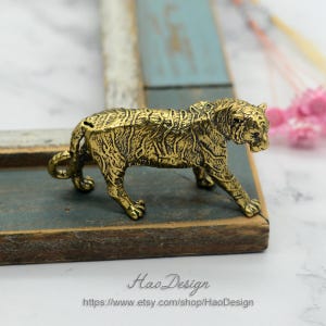 Pode incluir: Uma figurinha de tigre de metal dourado. O tigre est&aacute; em p&eacute; sobre uma superf&iacute;cie de madeira. A figurinha &eacute; detalhada com listras e um acabamento texturizado.