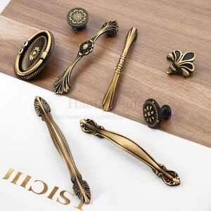 以下が含まれることがあります： アンティーク真鍮製のキャビネット金具の詰め合わせ。つまみや引き手など。装飾的な花柄や葉柄のデザインが特徴で、ヴィンテージ感があります。明るい木材の表面に配置され、その複雑なディテールと経年変化した仕上がりを際立たせています。