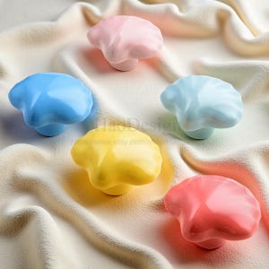 May include: A collection of five colorful ceramic cabinet knobs in shades of pink, blue, yellow, and red. Each knob has a unique, organic shape and sits on a short, rounded base. The knobs are arranged on a soft, textured surface.