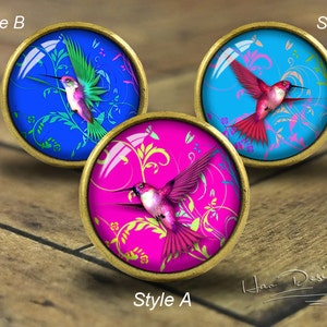 May include: Three round, gold-toned, decorative knobs with a glass top. Each knob features a different colored background with a hummingbird in flight. The backgrounds are blue, pink, and turquoise. The hummingbirds are green, pink, and red.