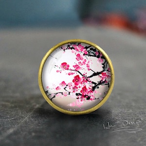 以下が含まれることがあります： 真鍮仕上げの丸いキャビネットノブ。ノブは、白い背景に黒い枝にピンクの桜の花の絵が描かれたガラスインサートを備えています。デザインには「Hao Design」の文字が含まれています。