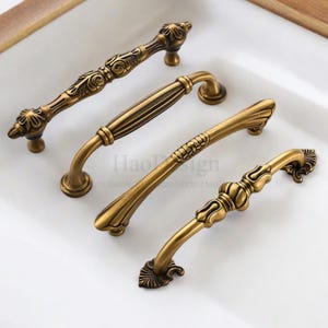 以下が含まれることがあります： 装飾的なデザインのアンティーク真鍮キャビネットハンドル4つ。ハンドルは形状と詳細が異なり、花柄や幾何学模様が特徴です。ハードウェアは白い表面に配置され、古びた真鍮仕上げが強調されています。