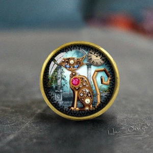 Könnte beinhalten: Eine goldfarbene Metallstiftspange mit einer Glaskuppel, die eine Katze im Steampunk-Stil mit Zahnrädern und einem Zifferblatt zeigt. Die Katze steht in einer blauen und grünen Landschaft mit Säulen und einer Sense.