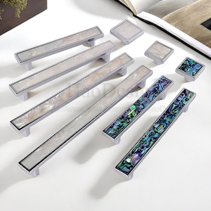 Może przedstawiać: Kolekcja okuć do szafek, w tym długie uchwyty i kwadratowe gałki. Uchwyty mają srebrne wykończenie z prostokątną inkrustacją z iryzującej masy perłowej. Niektóre uchwyty i gałki mają kolorową inkrustację ze skorupy abalonu.