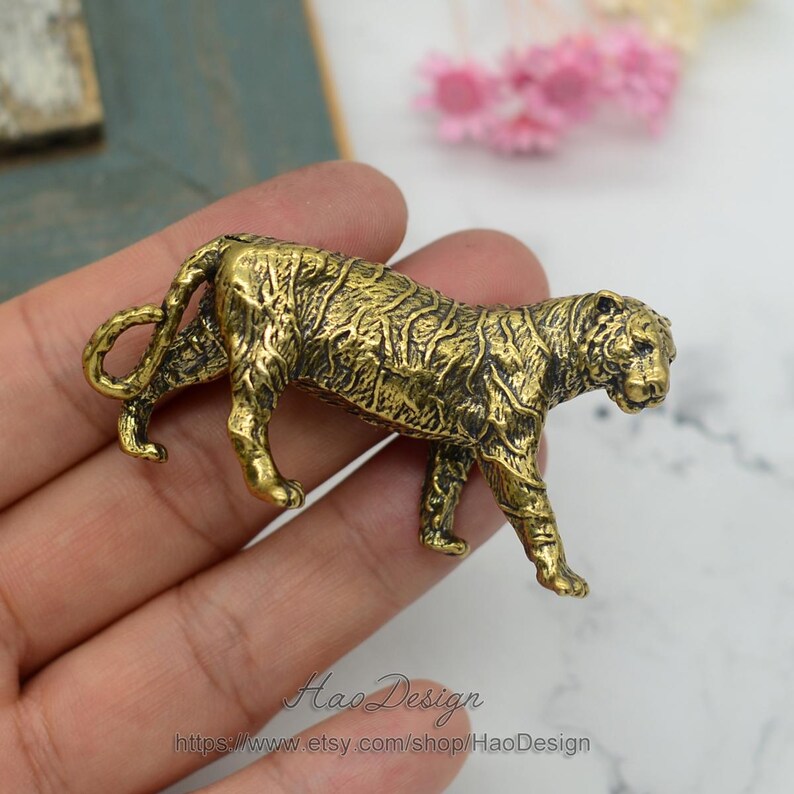 Pode incluir: Uma figurinha de tigre de metal dourado com marcas detalhadas. A figurinha est&aacute; em p&eacute; sobre uma m&atilde;o.