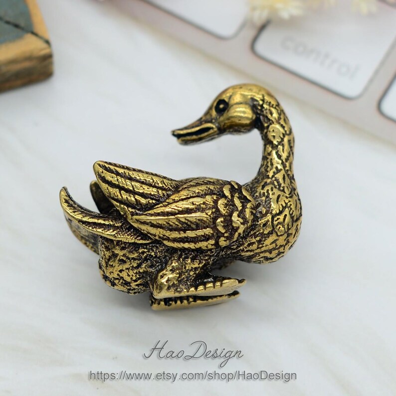 May include: A gold-coloured metal duck figurine with detailed feathers and a textured surface. The duck is in a sitting position with its head turned to the side.