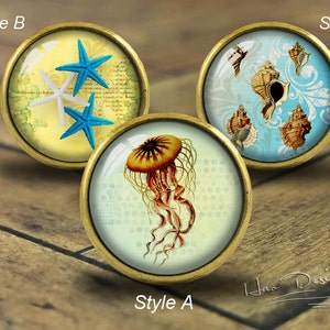 Puede incluir: Tres pomos redondos, de color dorado, decorativos para armarios con inserciones de vidrio. Los pomos presentan un tema náutico con imágenes de estrellas de mar, conchas marinas y una medusa. El texto "Style A", "Style B" y "Style C" está impreso debajo de cada pomo.