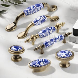 May include: A selection of antique brass cabinet handles and knobs. The handles have a white ceramic centre with a blue floral pattern. The knobs also feature the same design. The hardware is displayed on a white surface.